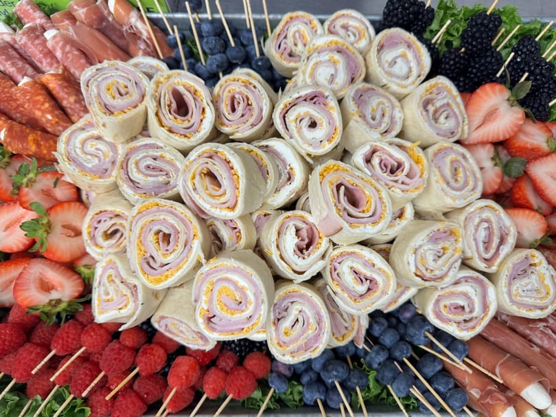 A platter of ham and cheese pinwheel wraps, with berries, strawberries, and cured meats.