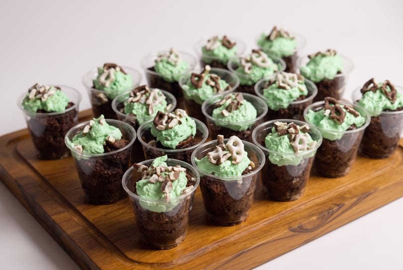 Mini chocolate desserts with green frosting and pretzel toppings on a wooden tray.